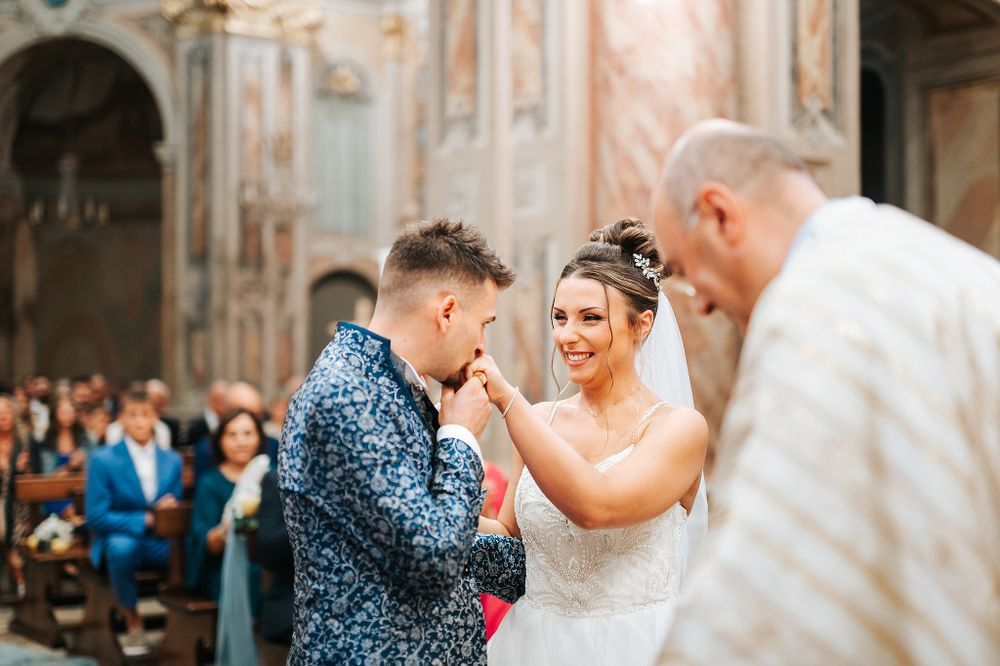 Groom kisses bride's hand tenderly during romantic wedding ceremony in ornate Italian church.