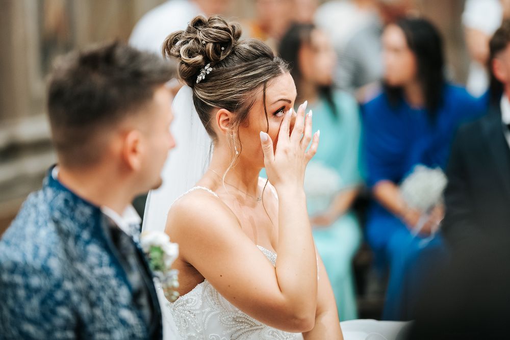 Emotional bride wipes tears during heartfelt wedding ceremony with guests in background.