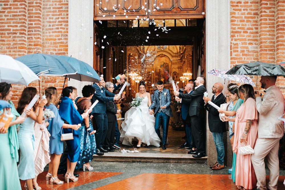 Newlyweds exit ornate church doors as guests celebrate with confetti and sparklers in rain.