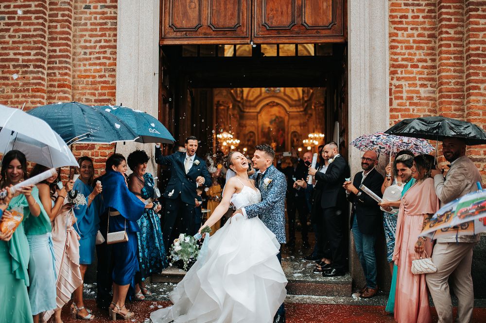 Joyful newlyweds walk out of church entrance as guests with umbrellas toss confetti in rain.