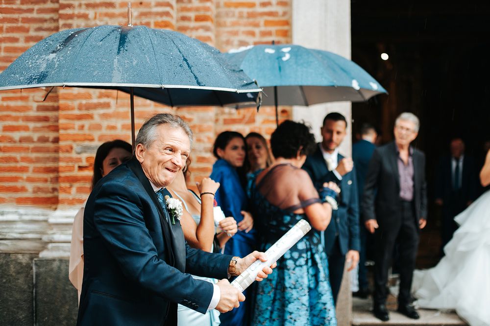 Wedding guests sheltering under umbrellas outside a brick church entrance on a rainy wedding day.