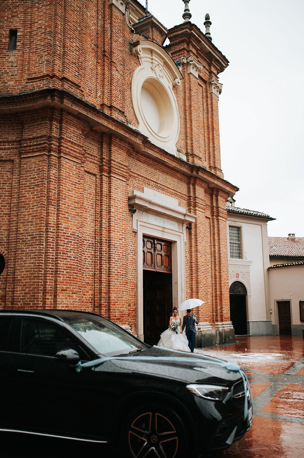 Bride and groom exit a historic brick church in the rain holding a white umbrella on their wedding day.