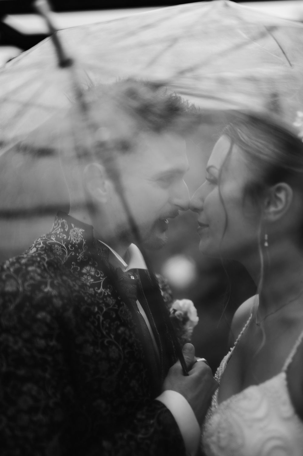 Black and white intimate moment of bride and groom sheltering under a transparent umbrella in the rain.
