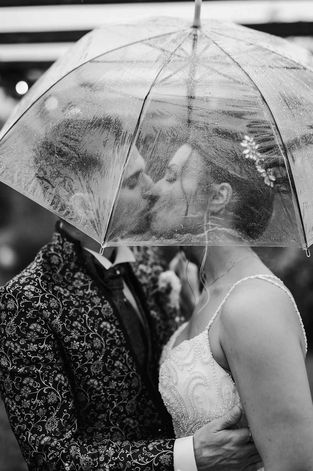 Bride and groom share a romantic kiss under a clear bubble umbrella on a rainy wedding day.