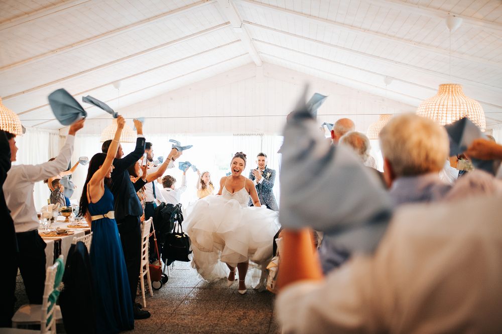 Joyful bride enters wedding reception as guests wave blue napkins in celebration inside a marquee.