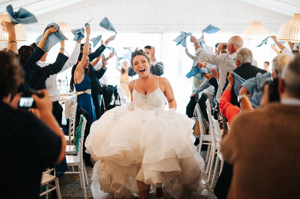 Beaming bride in a ballgown walks through cheering guests waving blue napkins at wedding reception.