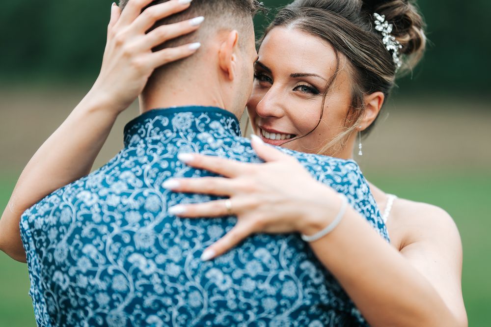 Smiling bride in a floral blue dress embraces her groom outdoors with a green nature backdrop.