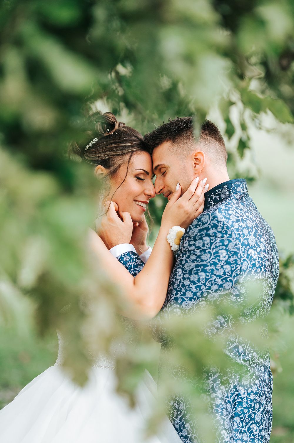 Bride and groom tenderly touching foreheads and smiling surrounded by lush green leaves outdoors.