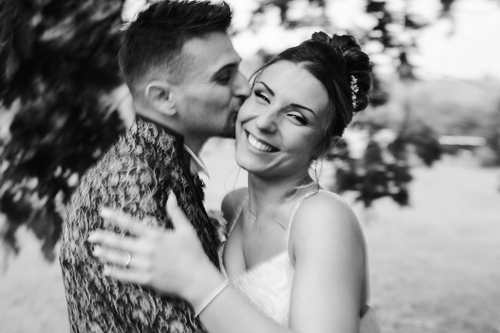 Black and white photo of groom kissing smiling bride on the cheek outdoors at wedding.
