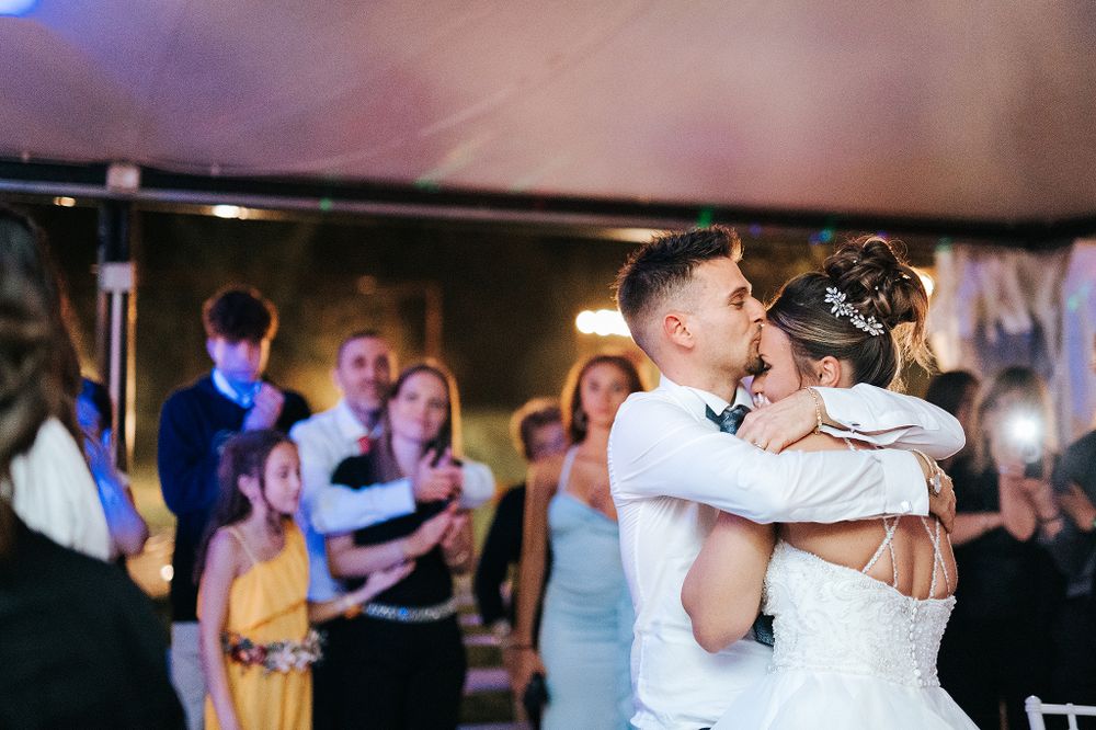 Bride and groom sharing their first dance embrace surrounded by smiling wedding guests in a tent.