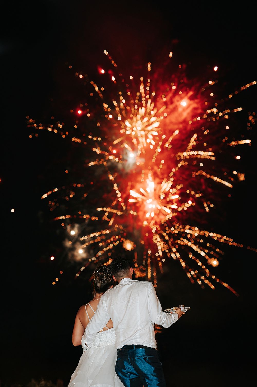 Couple watching vibrant red fireworks display together at their outdoor wedding celebration.