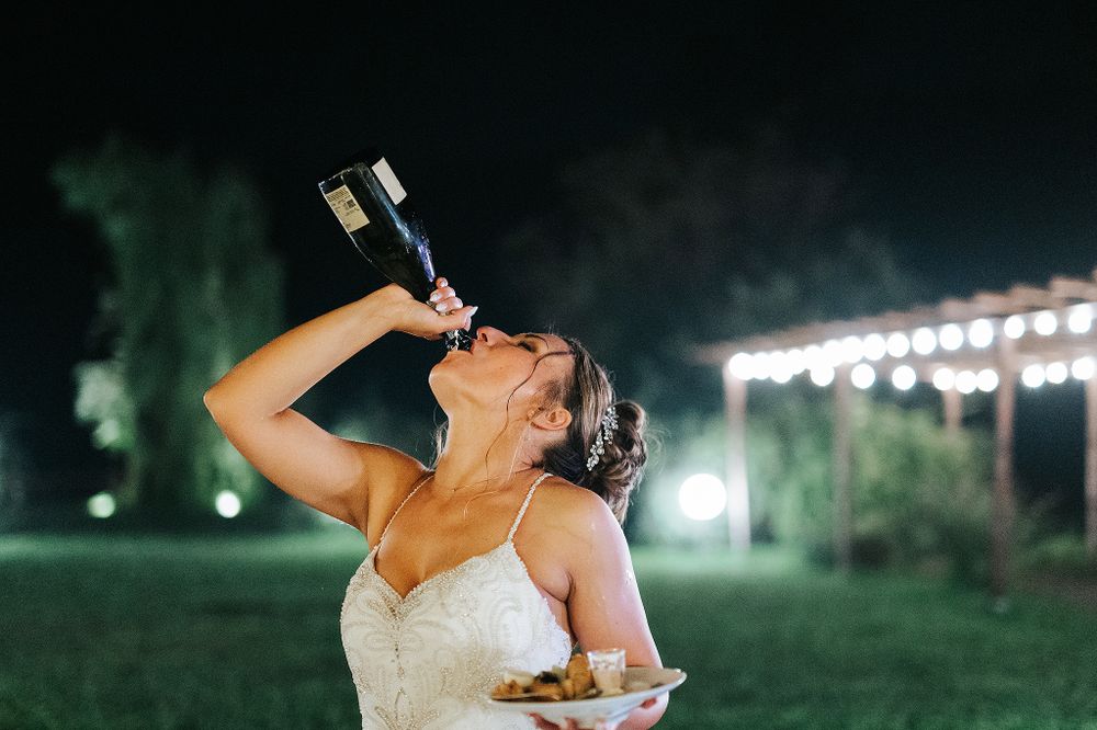 Joyful bride drinking champagne from bottle at outdoor evening wedding reception.
