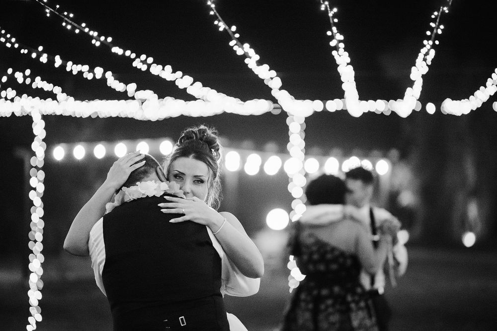 Two women sharing an emotional hug under string lights at wedding reception.