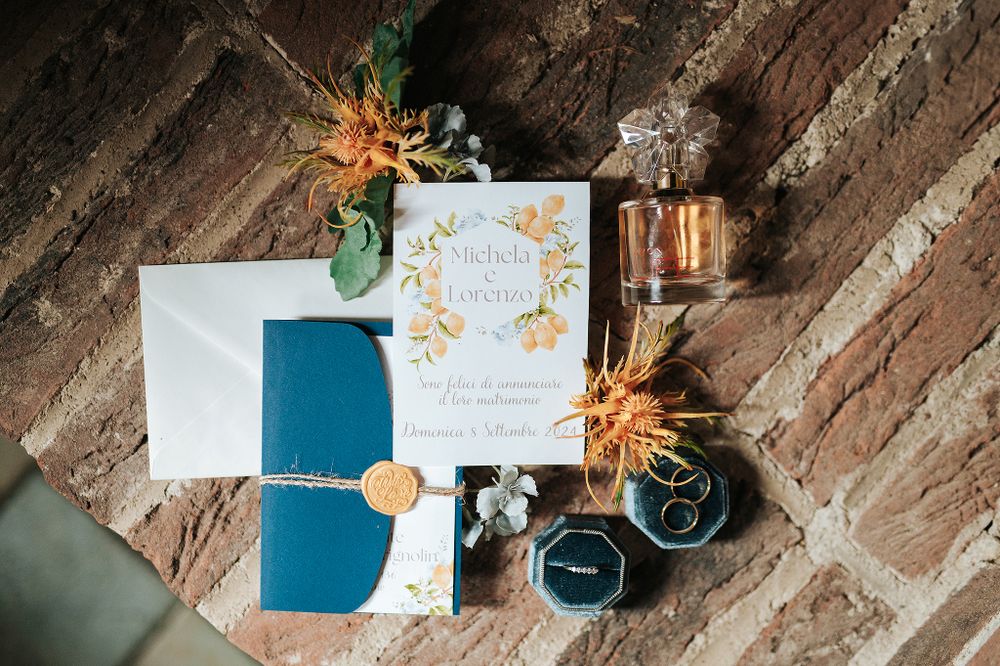 Elegant wedding stationery suite with navy blue envelope, floral invitation, wax seal, and ring boxes on rustic brick surface.
