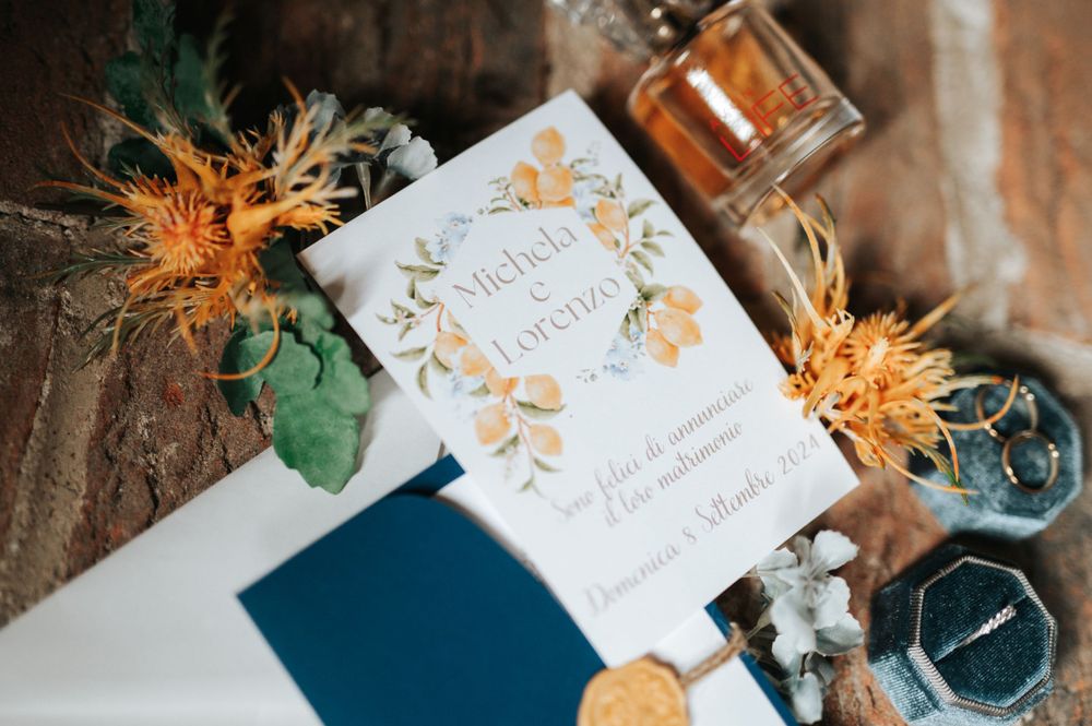 Close-up of floral wedding invitation with yellow botanical wreath design, navy envelope, and wax seal on brick background.