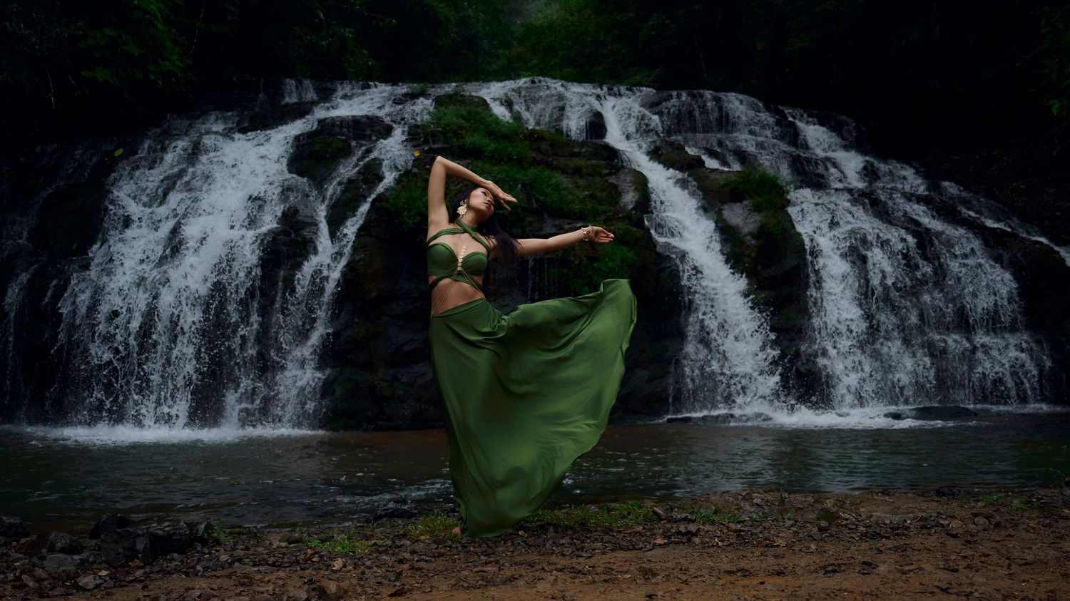 Elegant model in flowing emerald dress performs graceful pose against dramatic waterfall backdrop in nature.
