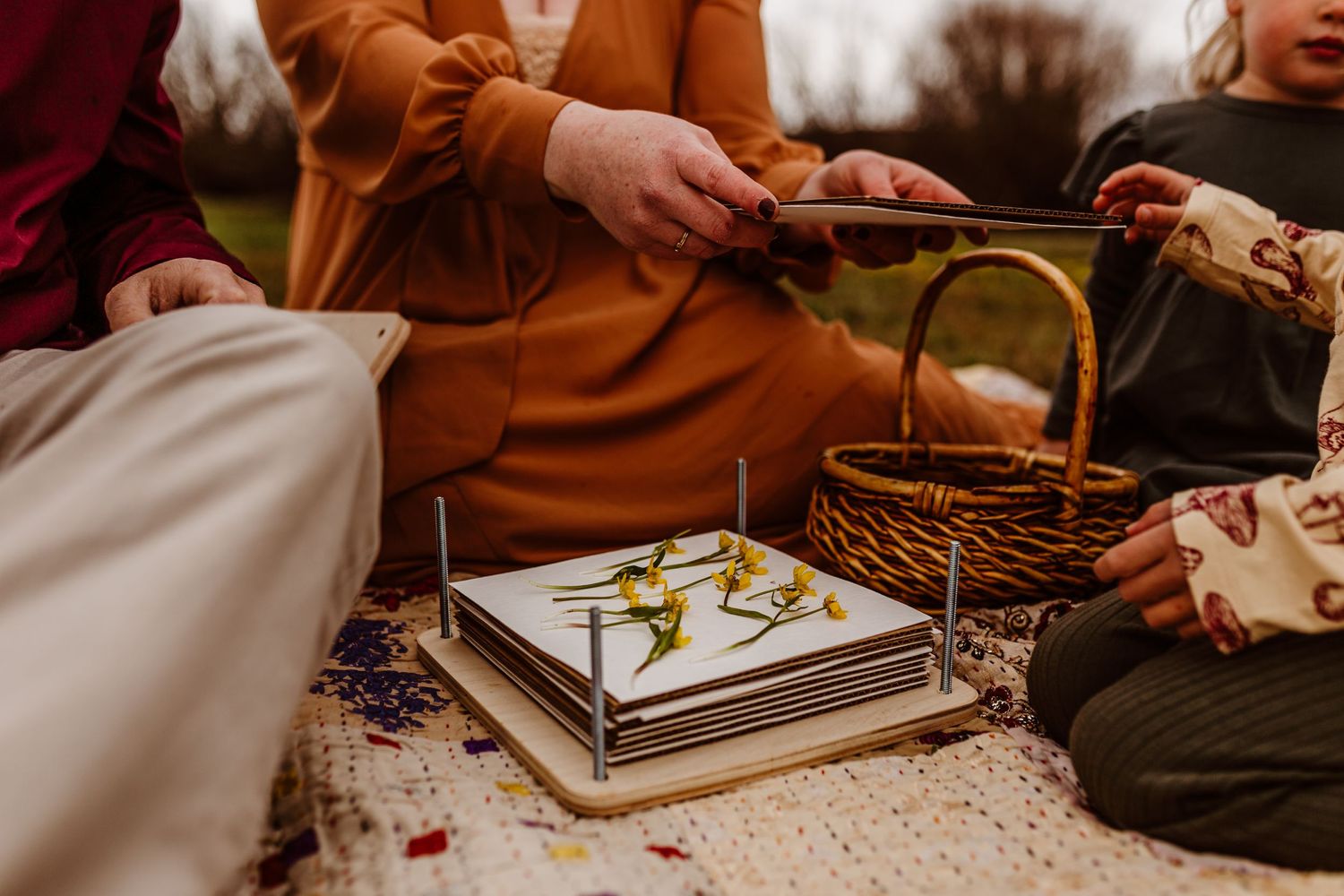 Family sitting outdoors on blanket with wicker basket and a flower press with fresh flowers.