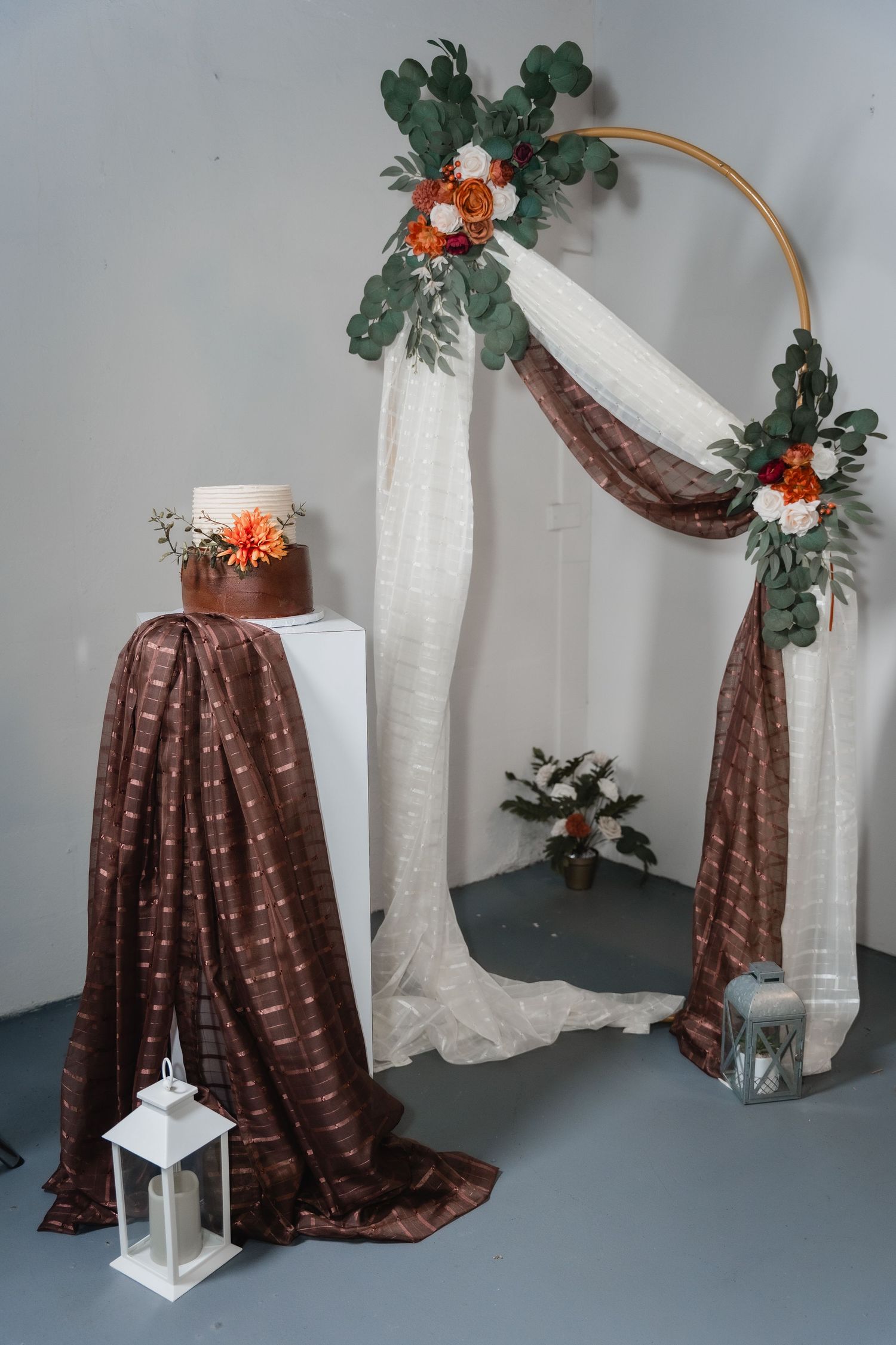 Wedding backdrop decor featuring a gold hoop arch with floral accents, brown draping fabric, and a white lantern accessory.
