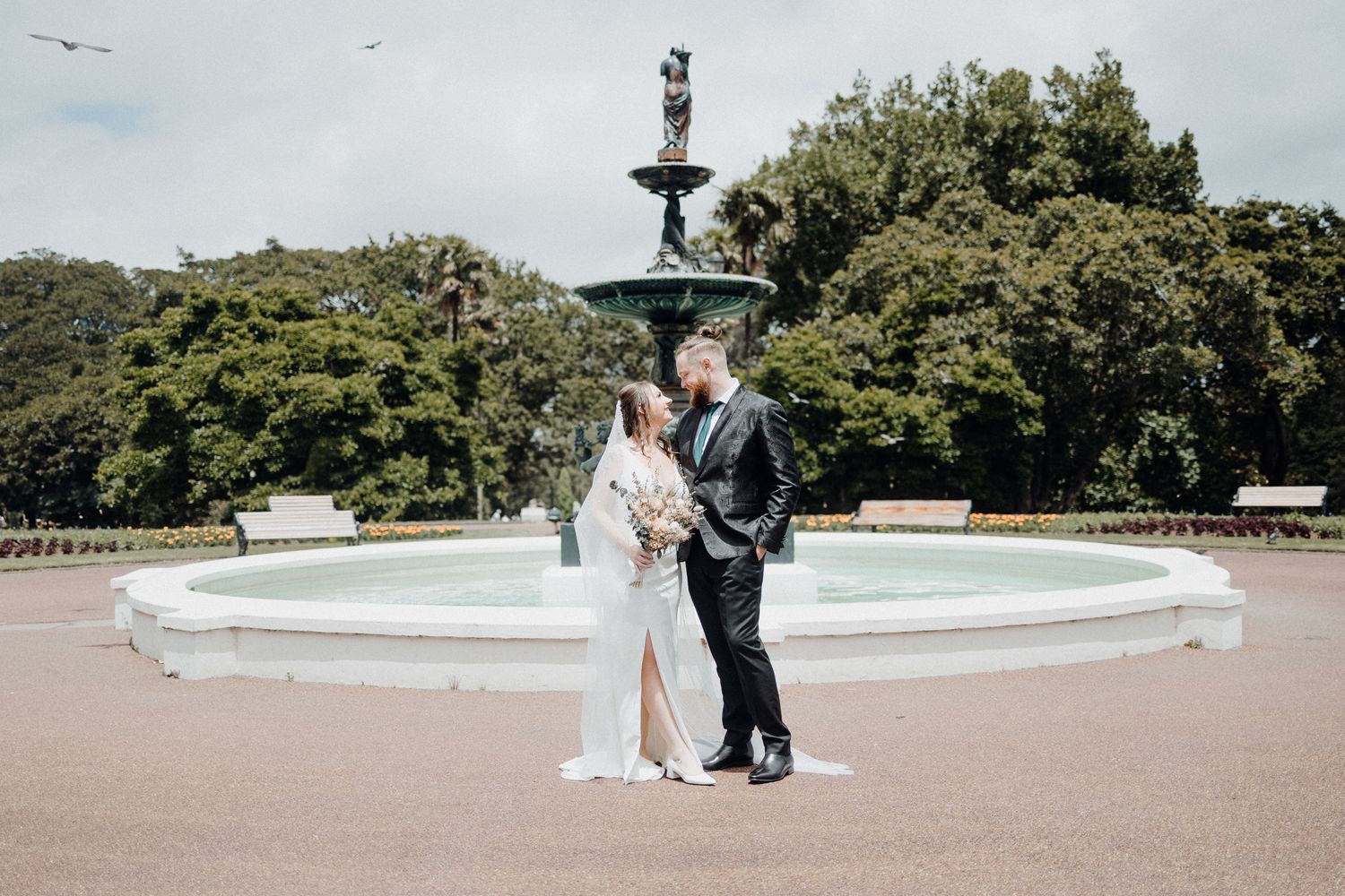 A Couple in front of an ornate garden fountain surrounded by lush green trees on a sunny day, captured by Apertured Media Auckland Wedding