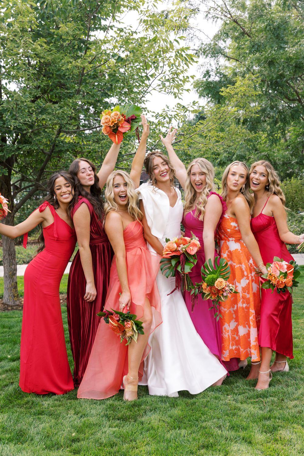Wedding party in coordinating red, coral, and pink dresses poses outdoors with bright orange floral bouquets on green lawn.
