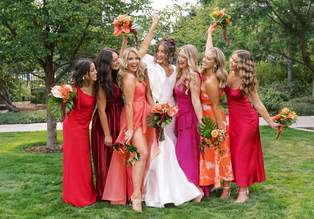 Bridesmaids in vibrant shades of red and pink dresses celebrate joyfully with bouquets in an outdoor garden setting.