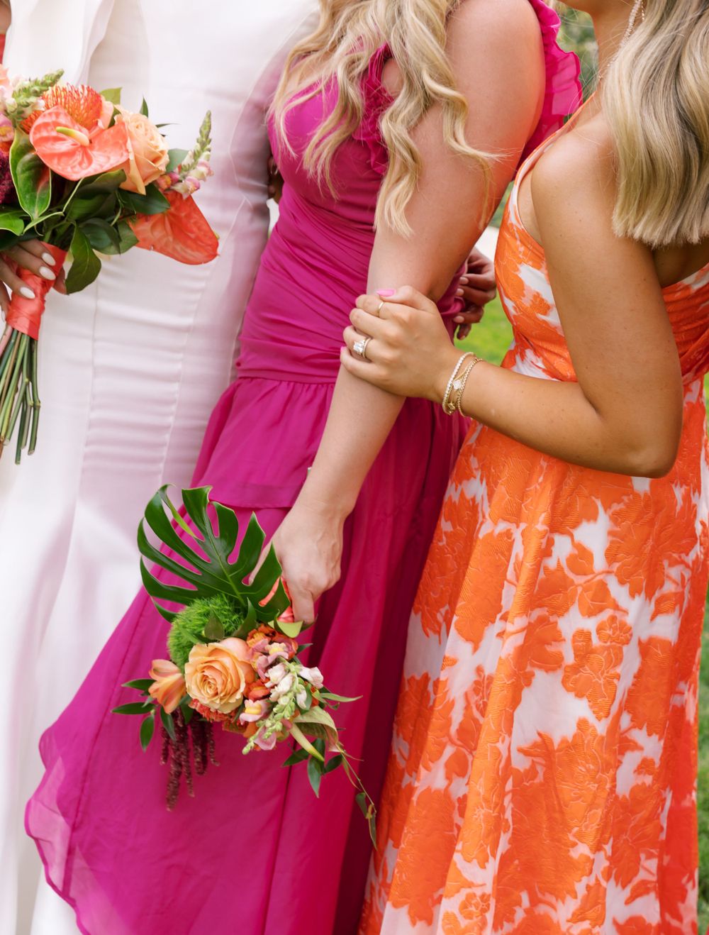 People in colorful bridesmaid dresses of magenta and orange hold tropical bouquets with peach roses and monstera leaves.