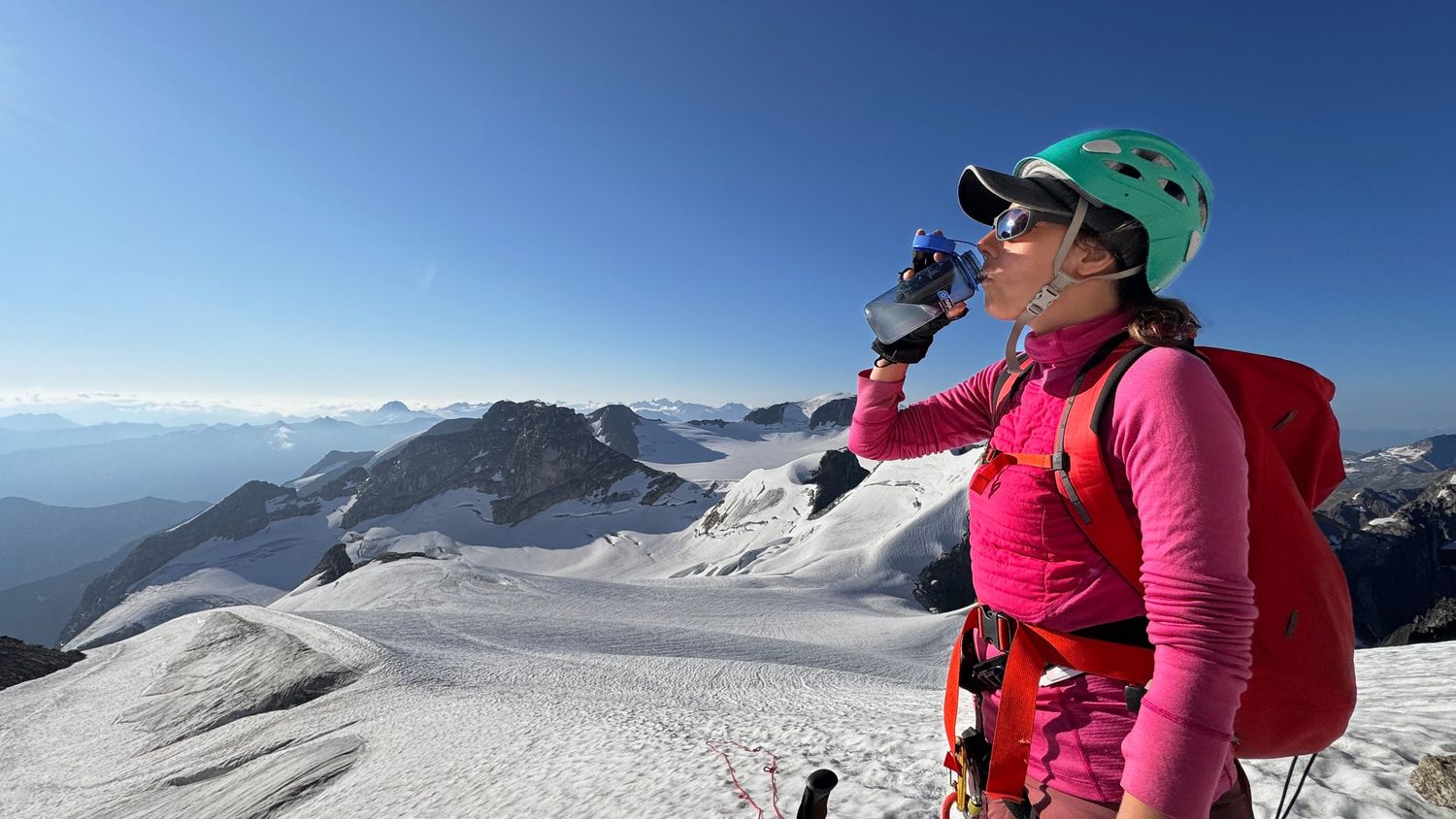 Mountain climber Kristen in a pink shirt and green helmet takes a drink while overlooking snowy alpine peaks and glaciers in the Bugaboos