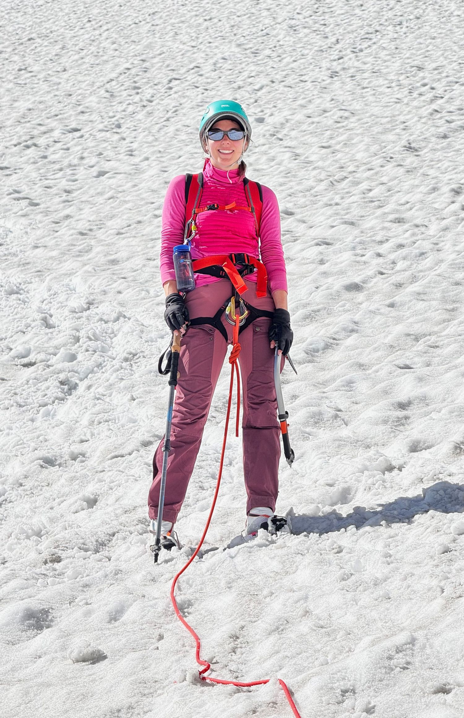 Kristen Lusk mountaineering in bright pink gear and climbing harness stands on a snowy glacier with ropes and safety equipment.