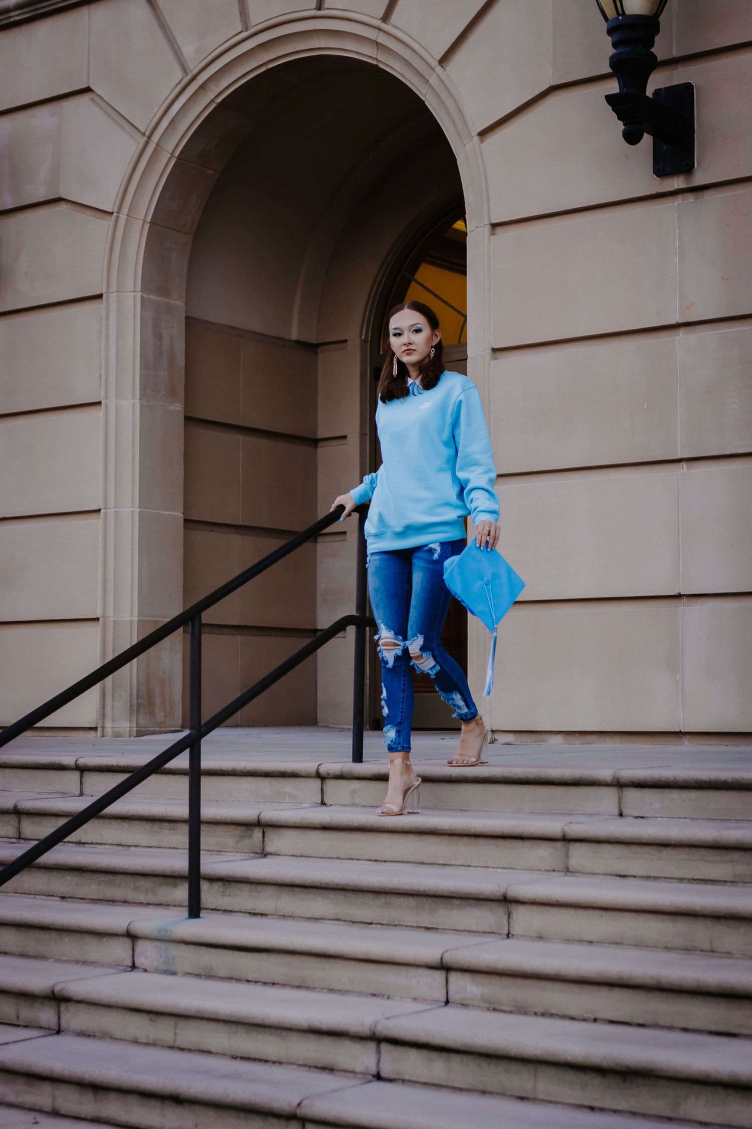 A person in a blue sweater and ripped jeans descends stone steps of a classic building.