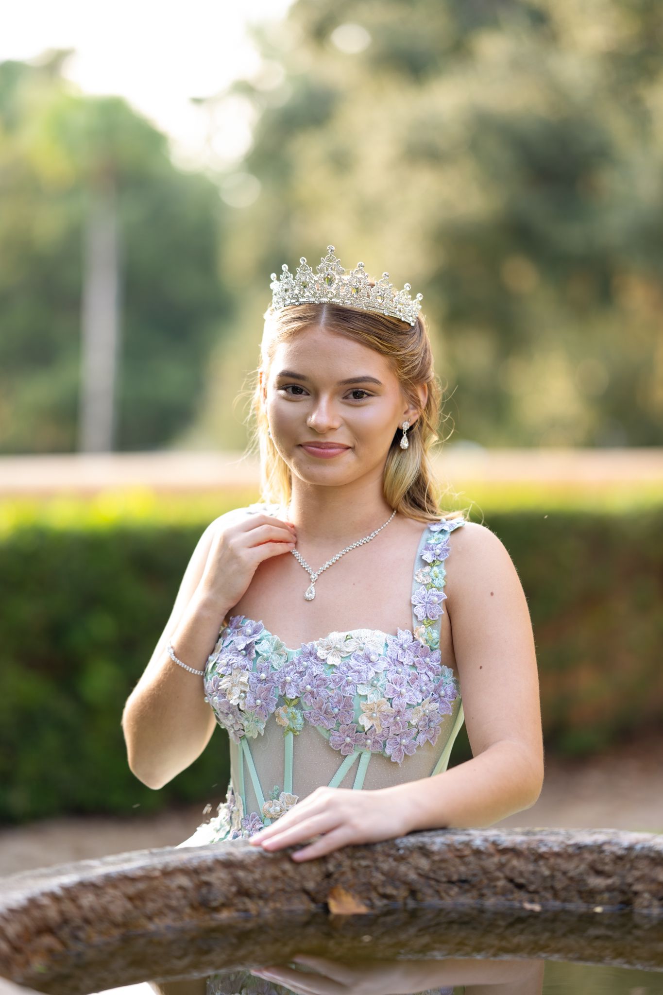 Young woman in a mint gown with floral details and tiara poses elegantly by a garden fixture.