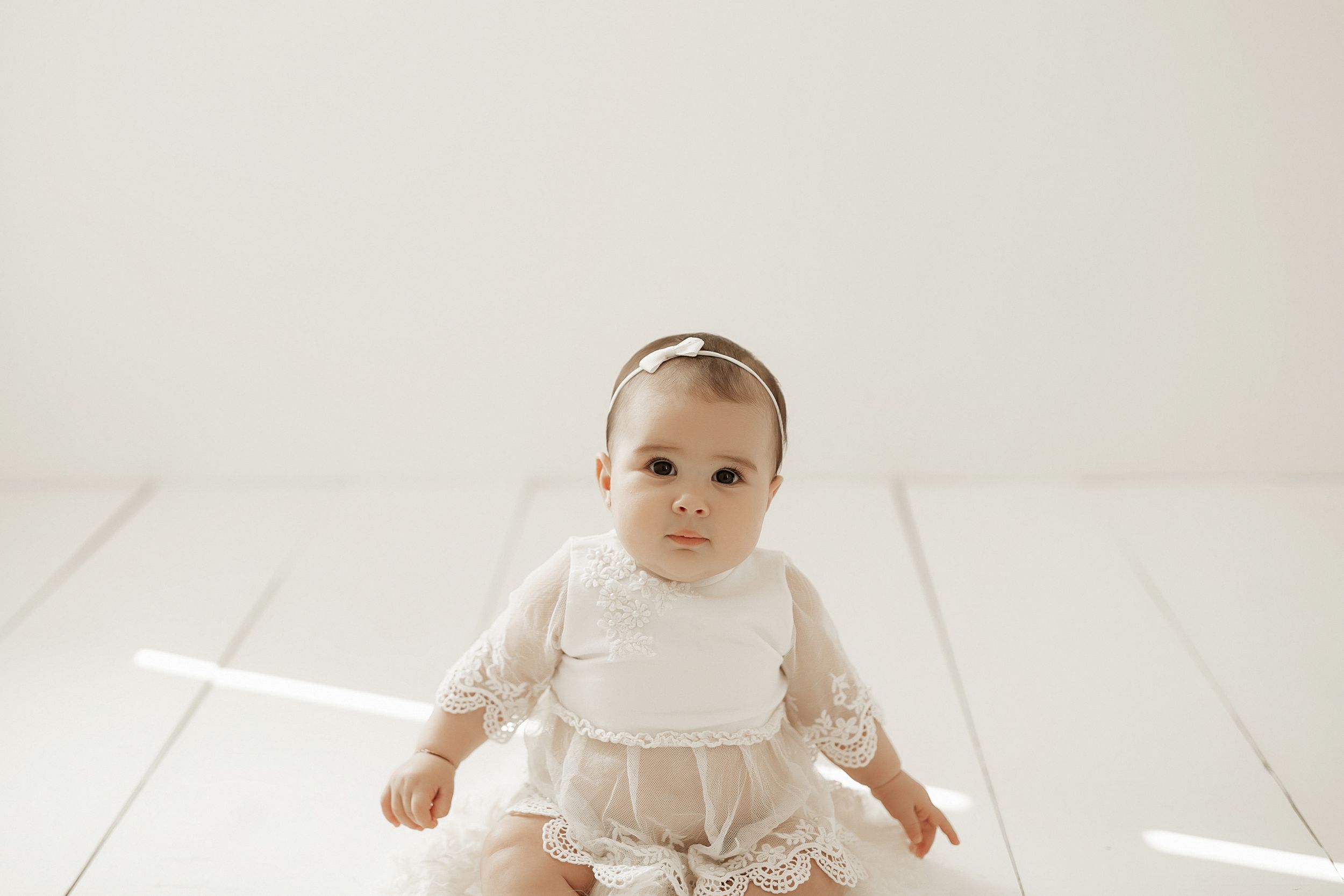 Baby in white lacy dress and bow headband sitting on white wooden floor.