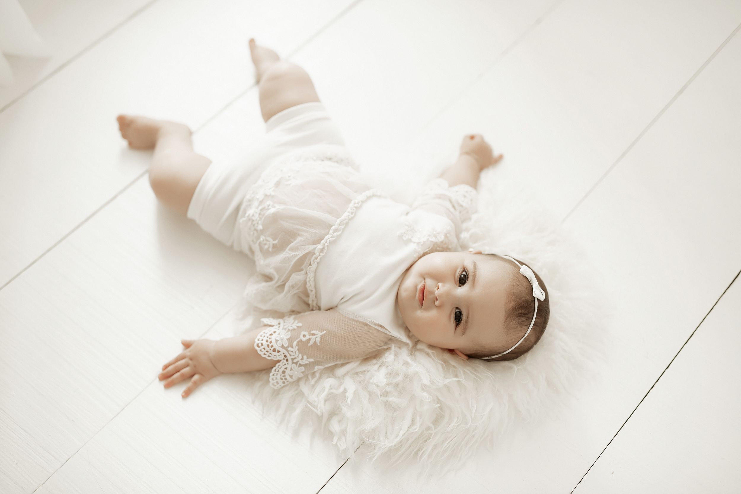 Baby in white lacy outfit lying on light wooden floor.