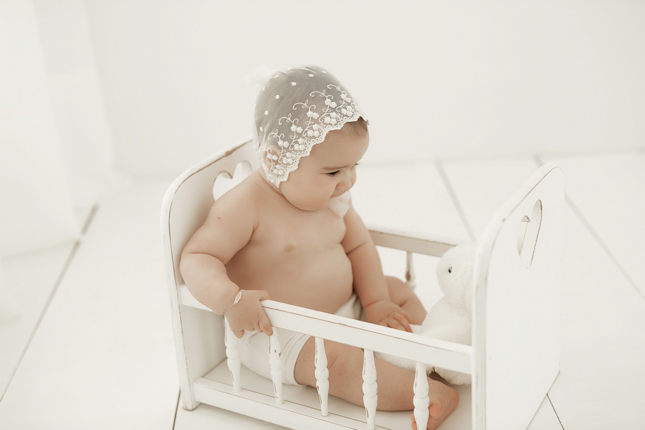 Baby wearing white lace bonnet sits in small white wooden doll crib against light backdrop.