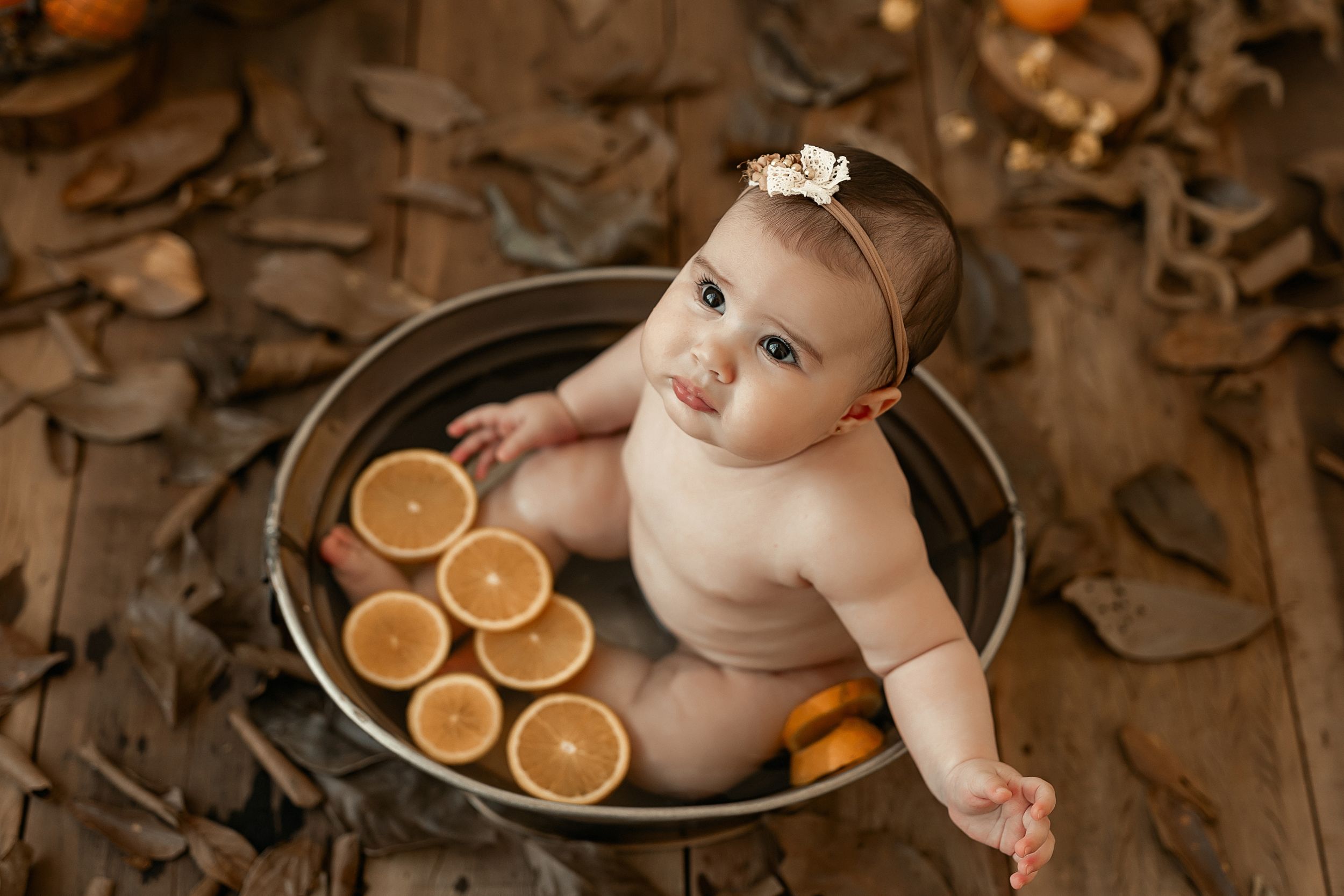 Baby in metal tub with sliced oranges and autumn decorations during themed photoshoot.