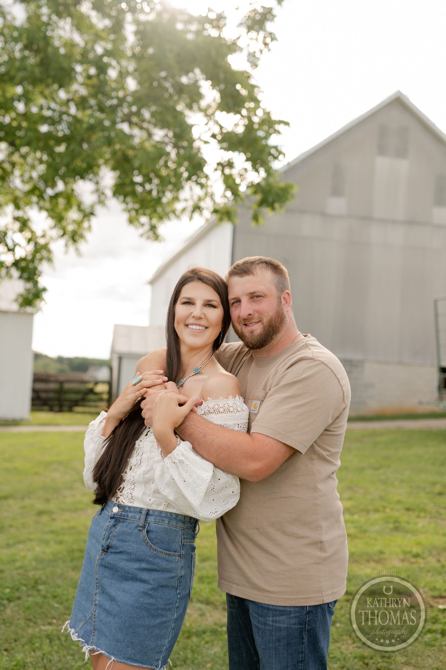 Couple embraces in casual attire against backdrop of rural barn and green trees.