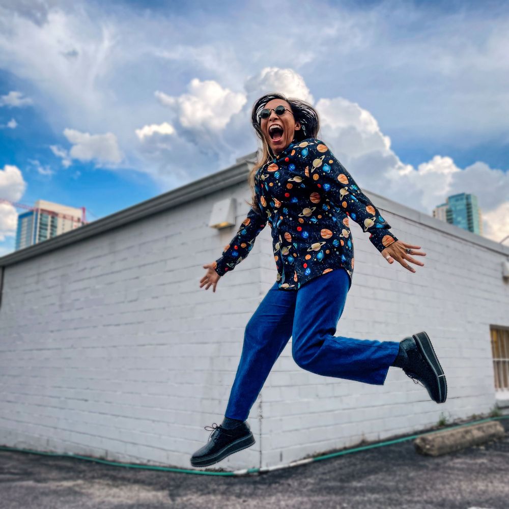 A person in a colorful planet patterned shirt and blue pants joyfully leaps in front of a white wall on a sunny day.