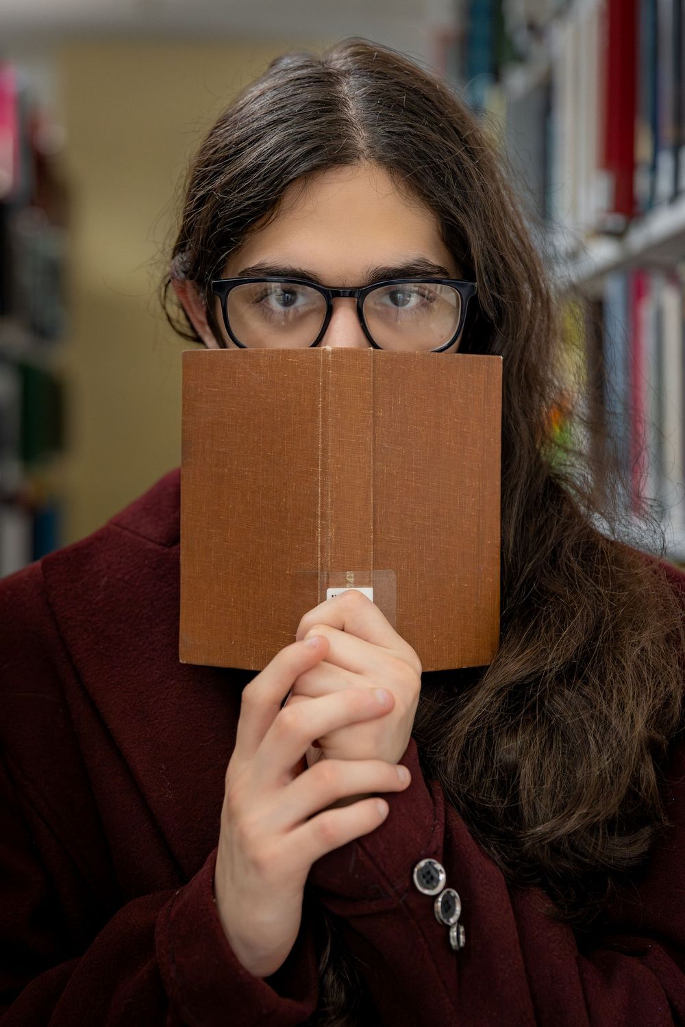 A young person with long hair and glasses holds an open book over their face in a library.