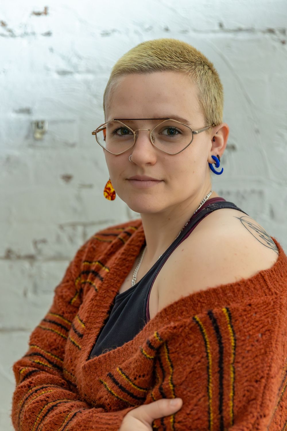 Young person with short blonde hair, glasses, colorful earrings, and rust orange cardigan poses against white brick wall.