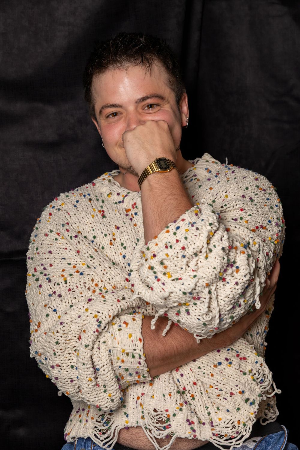 Man in a colorful speckled knit sweater laughing with hand over mouth, wearing a gold watch.