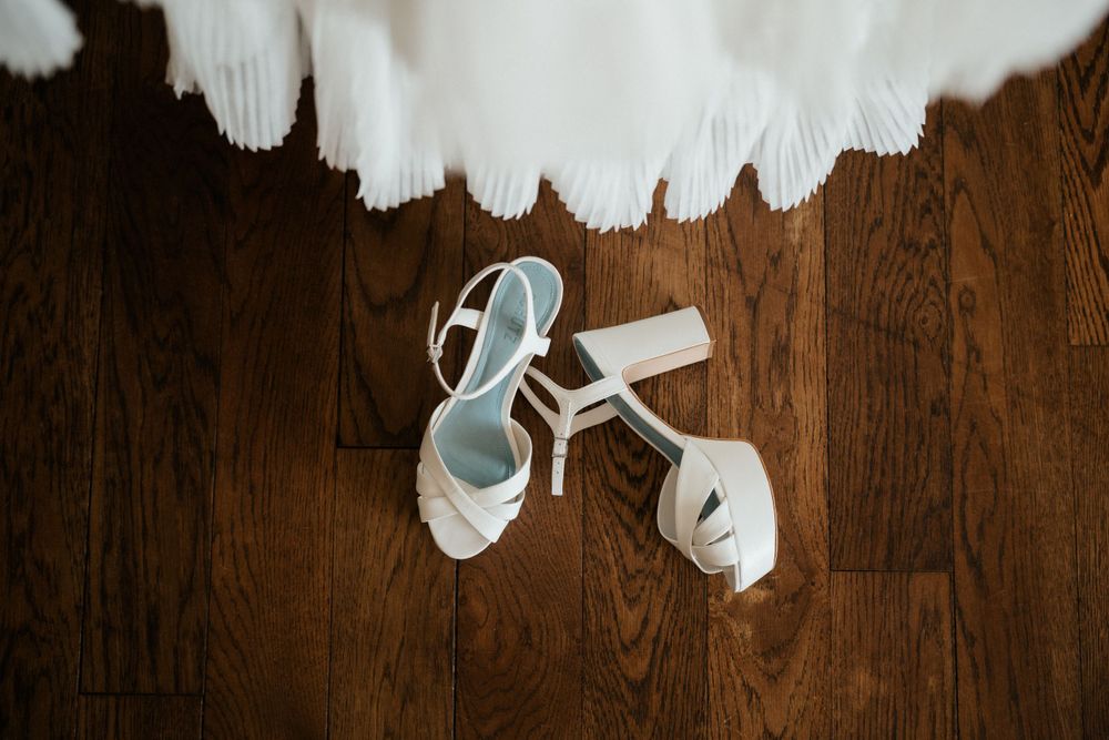 White bridal shoes placed on dark hardwood flooring next to flowing fabric.