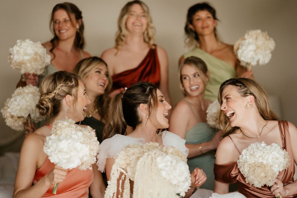 Bridesmaids in mixed-color dresses share laughter while holding white bouquets.