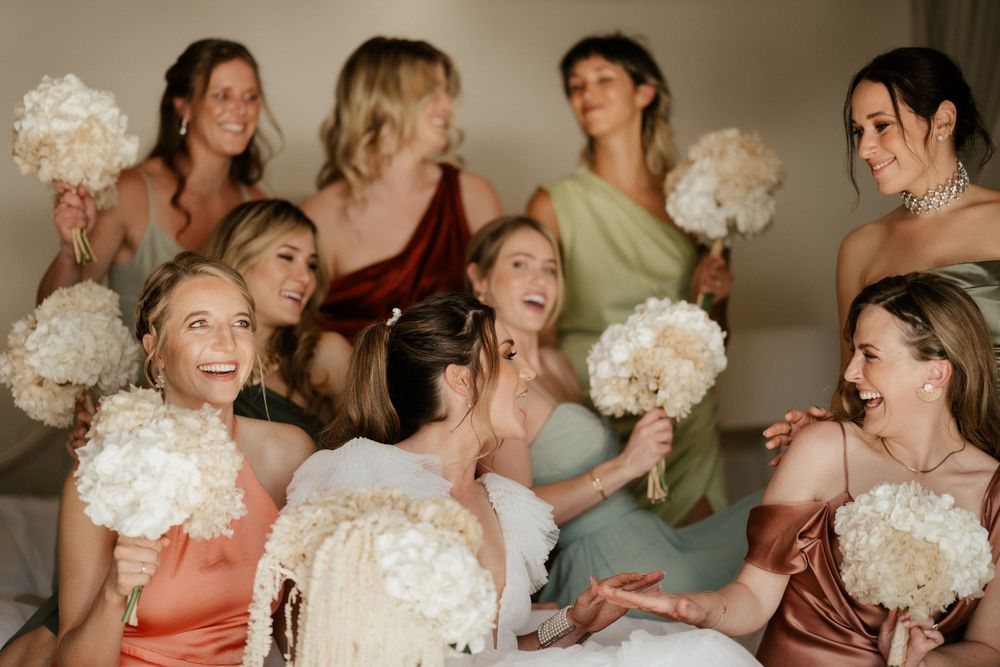Candid moment of a wedding party laughing and holding white bouquets in a warm-lit room.