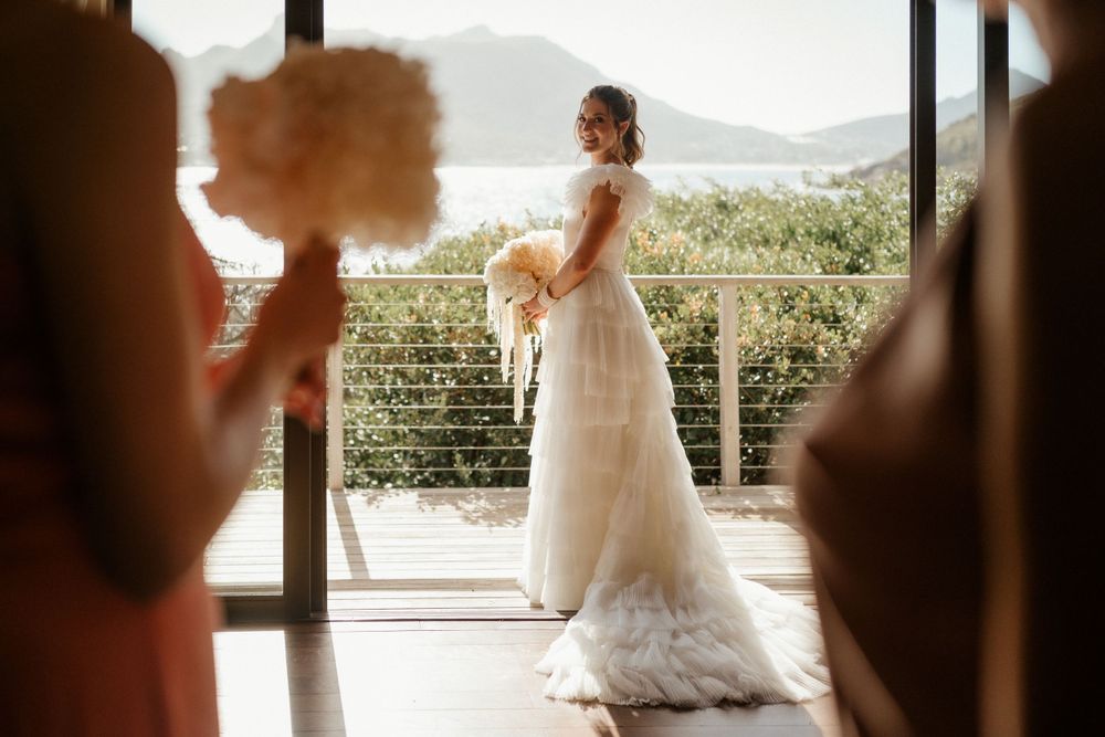 A bride in a flowing white gown poses by large windows with mountain and ocean views.