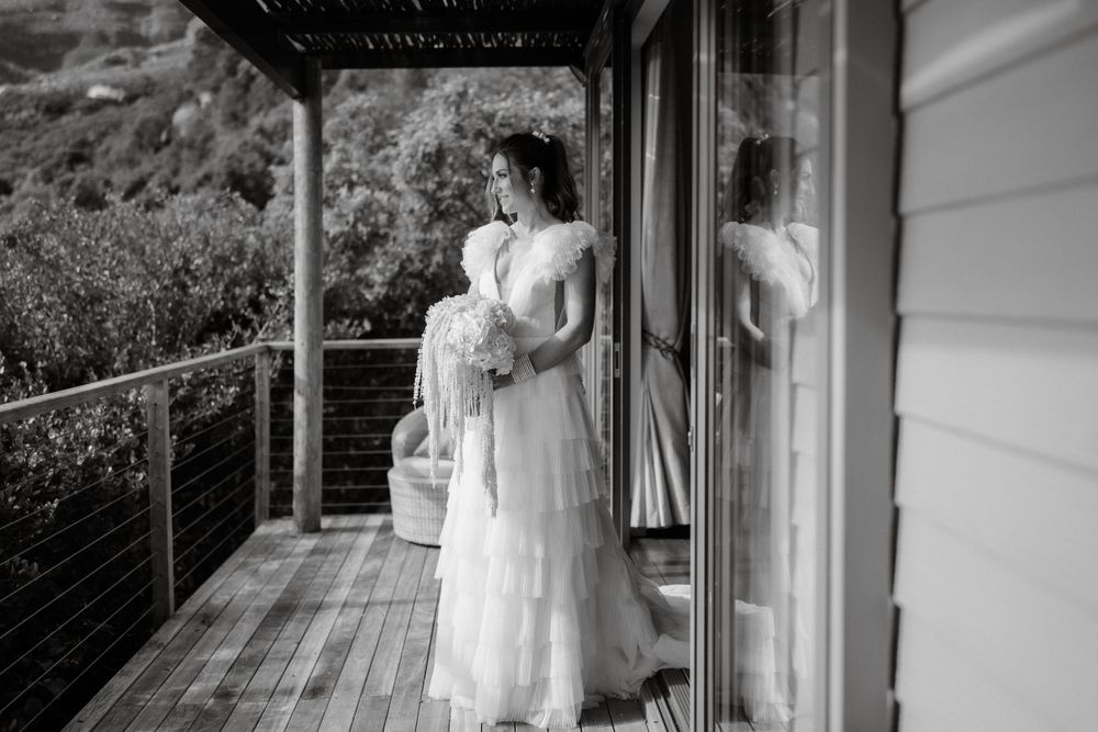 Elegant figure in flowing white dress stands on a modern deck with mountain views.