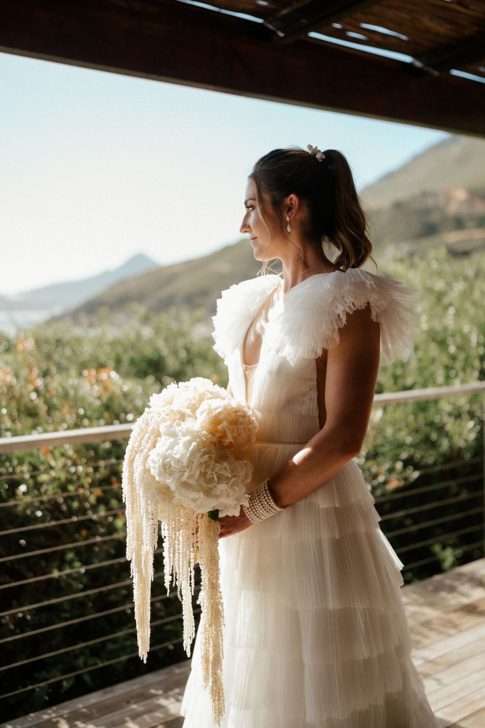 A bride in a layered tulle gown holds a cascading cream-colored bouquet while standing on a balcony with mountain views.