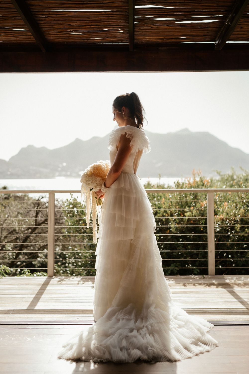 A white wedding gown silhouetted against mountains on a sunlit balcony.