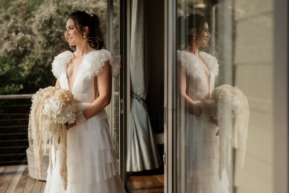 Elegant bride in white wedding dress holding dried floral bouquet poses next to reflective window.
