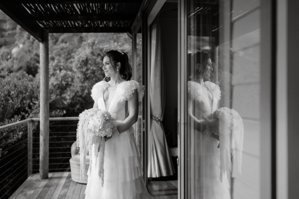Black and white photo of bride in lace dress standing on porch with reflection in window.