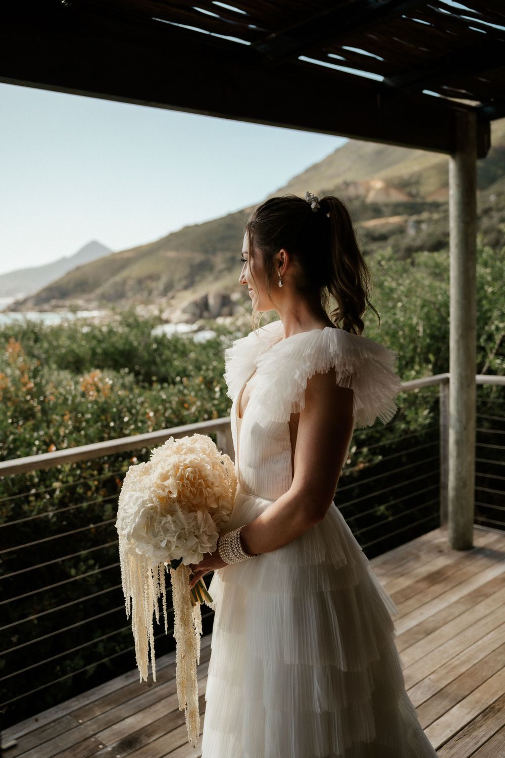 A bride in a flowing white dress holds a bouquet while standing on a deck overlooking mountain scenery.