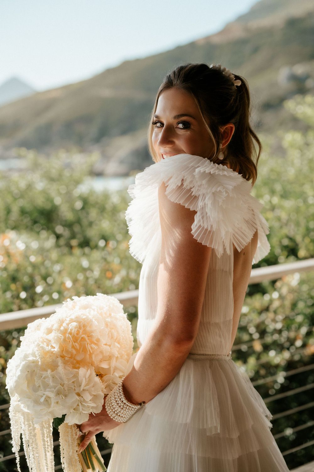 Bride in a romantic ruffled wedding dress holds a peach-colored bouquet against a mountainous landscape background.